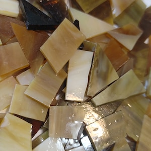 May include: A collection of irregularly shaped glass tiles in shades of brown, beige, and cream. The tiles vary in size and shape, with some appearing translucent. The overall effect is a mix of warm, earthy tones, suitable for mosaic art or craft projects.