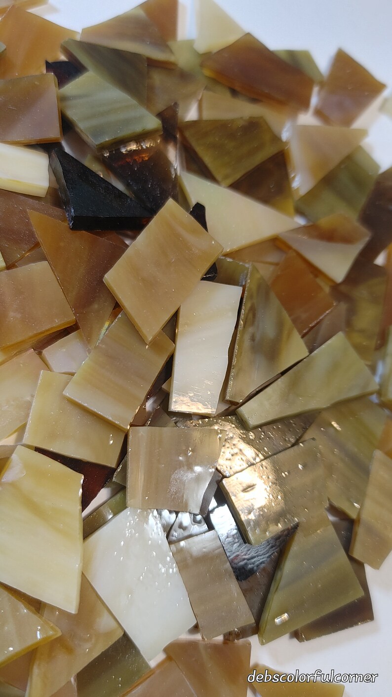 May include: A collection of irregularly shaped glass tiles in shades of brown, beige, and cream. The tiles vary in size and shape, with some appearing translucent. The overall effect is a mix of warm, earthy tones, suitable for mosaic art or craft projects.
