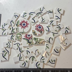 May include: A collection of white ceramic mosaic tiles featuring floral patterns with pink petals, green leaves, and dark blue stems. The tiles are arranged above a black ruler measuring 9 inches, showing various shapes including two circular pieces.