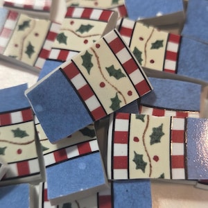 May include: A collection of small, rectangular tiles with a festive design. Each tile features a light beige background with red and white stripes, green holly leaves, and a blue border. The tiles are scattered.