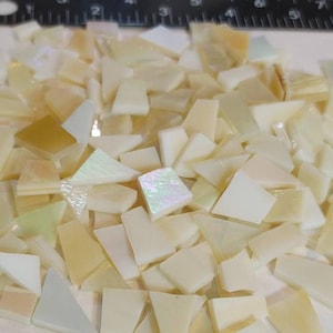 May include: A pile of small, irregular, white and cream-colored iridescent glass tiles. The tiles are various shapes and sizes, with some having a pearlescent finish.