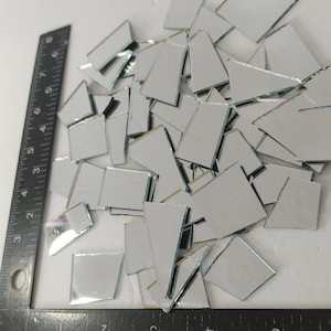 May include: A collection of small, irregularly shaped mirror tiles scattered on a white surface. A black ruler with inch measurements is positioned on the left side of the image, providing scale. The tiles vary in size and shape.