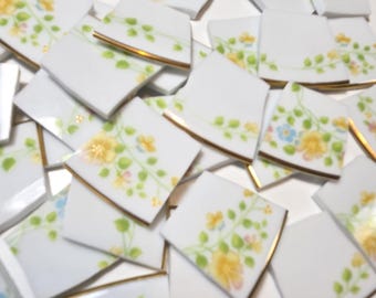 May include: A collection of white ceramic mosaic tiles featuring a floral pattern with yellow and blue flowers, green vines, and gold-trimmed edges. A black ruler is visible at the bottom, showing measurements in inches for scale.