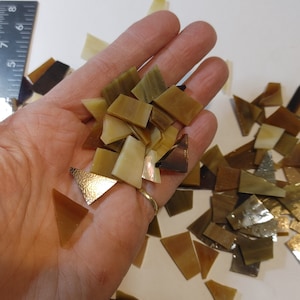 May include: A collection of small, irregularly shaped mosaic tiles in shades of brown, tan, and gold. The tiles are held in a hand, with some scattered on a white surface. The tiles vary in size and shape, with some having a textured, metallic appearance.