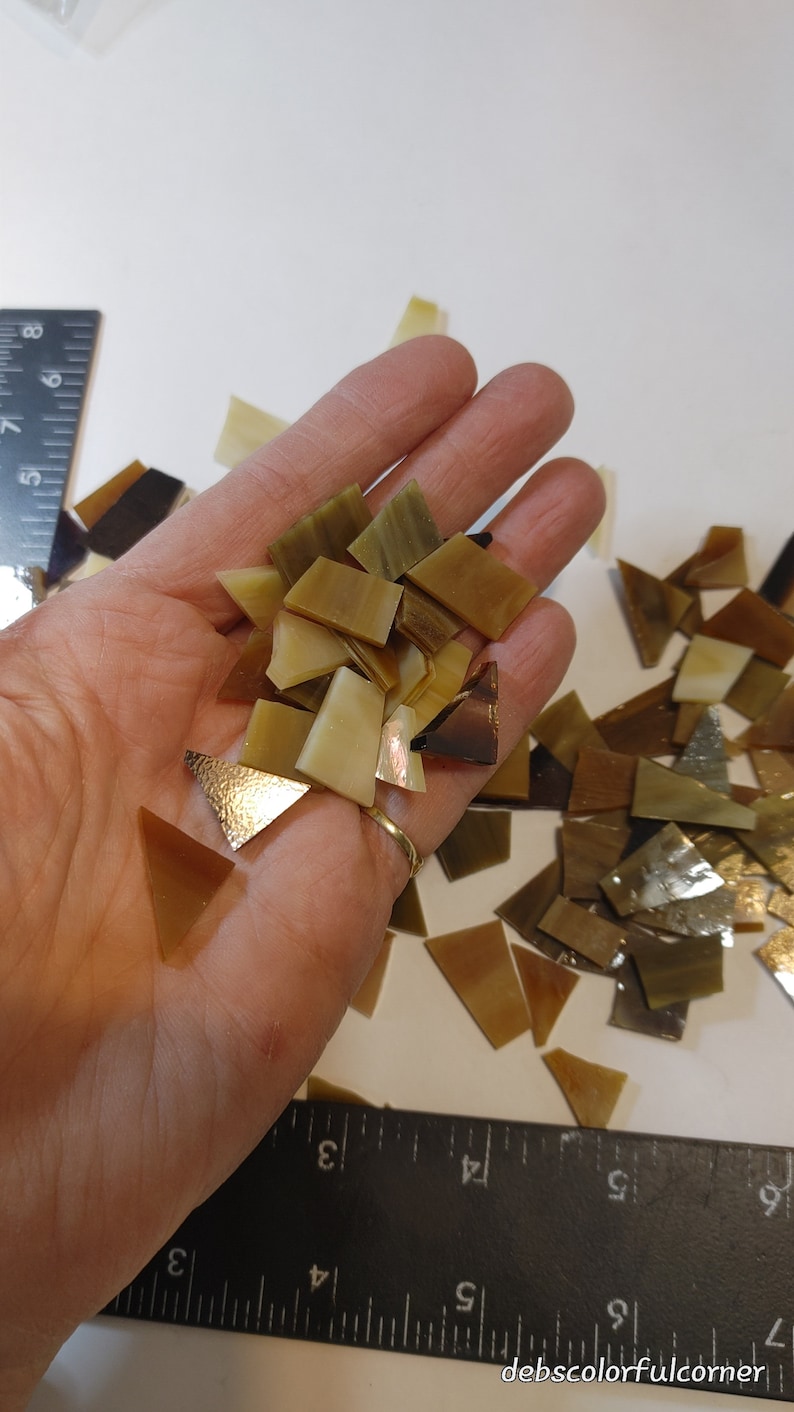 May include: A collection of small, irregularly shaped mosaic tiles in shades of brown, tan, and gold. The tiles are held in a hand, with some scattered on a white surface. The tiles vary in size and shape, with some having a textured, metallic appearance.