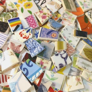 May include: A collection of colorful ceramic tile pieces with various patterns and designs. The tiles feature floral, geometric, and abstract motifs in a range of colors, including blue, pink, yellow, and orange. Some tiles have text. The pieces are irregularly shaped.