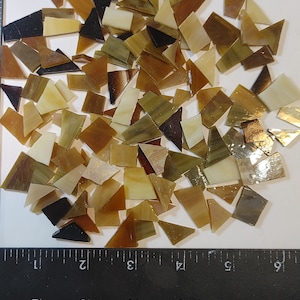 May include: A collection of irregularly shaped mosaic tiles in shades of brown, beige, and black. The tiles are scattered on a white surface, with a ruler providing scale. The tiles vary in size and shape, suitable for craft projects.