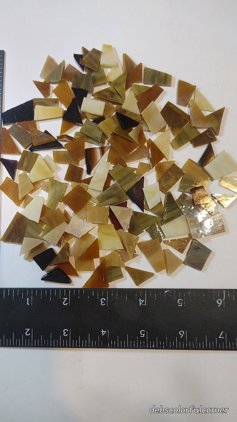 May include: A collection of irregularly shaped mosaic tiles in shades of brown, beige, and black. The tiles are scattered on a white surface, with a ruler providing scale. The tiles vary in size and shape, suitable for craft projects.
