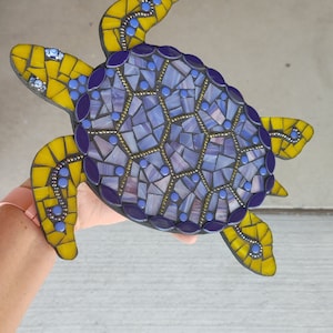 May include: A mosaic turtle sculpture with a purple and blue shell and yellow flippers. The turtle is made of small pieces of glass and has a textured surface.