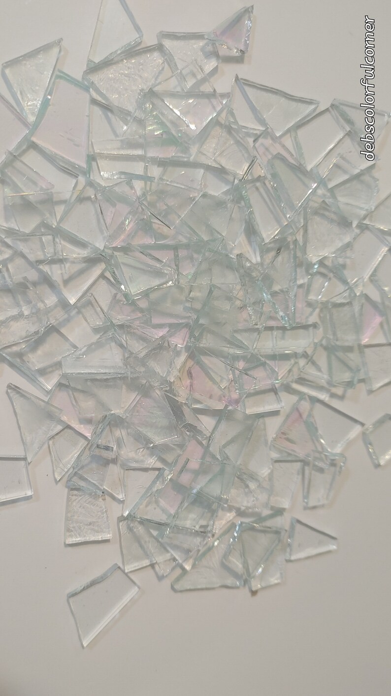 Puede incluir: Una colecci&oacute;n de fragmentos de vidrio transparente de forma irregular, con superficies iridiscentes. Las piezas est&aacute;n dispersas sobre un fondo blanco, creando una exhibici&oacute;n texturizada y reflectante. Los fragmentos var&iacute;an en tama&ntilde;o y forma, capturando la luz para producir un sutil efecto arco&iacute;ris.