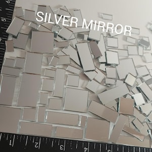 May include: A pile of small, irregular shaped pieces of silver mirror. The pieces are various sizes and shapes, and are arranged randomly. A ruler is visible at the bottom of the image, showing the size of the pieces.