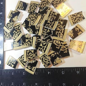 May include: A collection of small, rectangular tiles with a black background and gold floral patterns. The tiles are scattered across a white surface, with a ruler providing scale. The tiles have a decorative, vintage aesthetic.