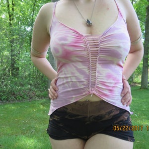 May include: A woman wearing a pink and white tie-dye tank top with a crocheted detail down the front. The tank top is paired with black shorts with a tie-dye pattern. The image was taken outdoors in a grassy area.