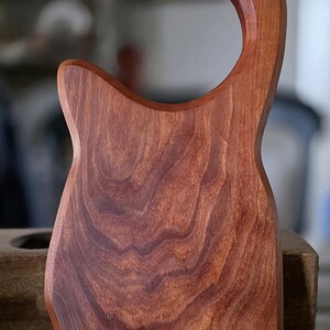 May include: A handcrafted wooden gua sha tool with a curved handle. The tool is made of reddish-brown wood, featuring visible wood grain patterns. The text "5D LEATHER BELLEVILLE TX" is visible on the bottom.