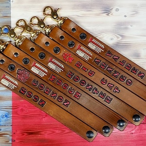 May include: Seven brown leather keychains with gold key rings and black snap closures. Each keychain has a red and white embroidered firefighter emblem and a name: "BOOC", "NOON", "MOOC", "HI BOOC", "BIG CAT", "CAT ARANJO", "VALDES".