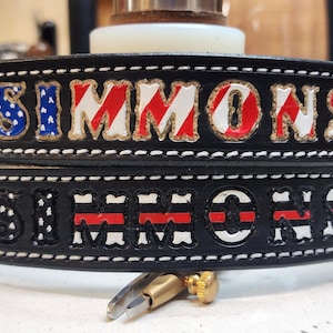 Pode incluir: Dois cintos de couro preto com costuras brancas. O cinto superior tem o nome "SIMMONS" escrito em letras brancas com listras vermelhas e azuis. O cinto inferior tem o nome "SIMMONS" escrito em letras brancas com listras vermelhas e brancas.