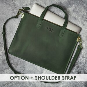 K&ouml;nnte beinhalten: Eine dunkelgr&uuml;ne Ledertasche f&uuml;r Laptops mit einem Schultergurt und einem Rei&szlig;verschluss. Die Tasche hat ein Monogramm mit dem Initial "GM" auf der Vorderseite.