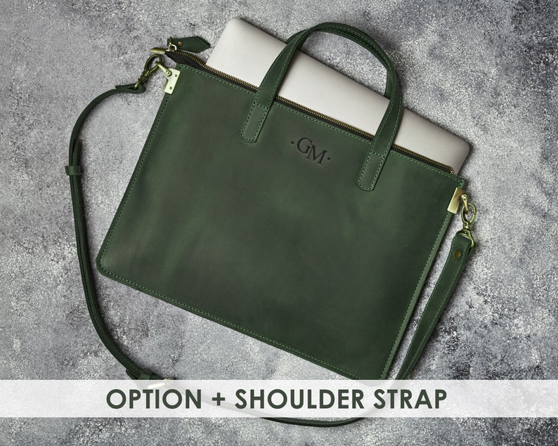 K&ouml;nnte beinhalten: Eine dunkelgr&uuml;ne Ledertasche f&uuml;r Laptops mit einem Schultergurt und einem Rei&szlig;verschluss. Die Tasche hat ein Monogramm mit dem Initial "GM" auf der Vorderseite.