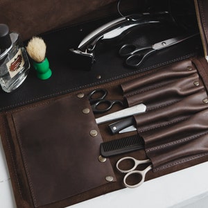 Op de afbeelding: Open bruin leren barbierkit met een zwart interieur. De kit bevat een flesje parfum, een scheerkwast, een elektrische tondeuse, een schaar, een recht scheermes, een kam en andere verzorgingsgereedschappen. Op een wit oppervlak.