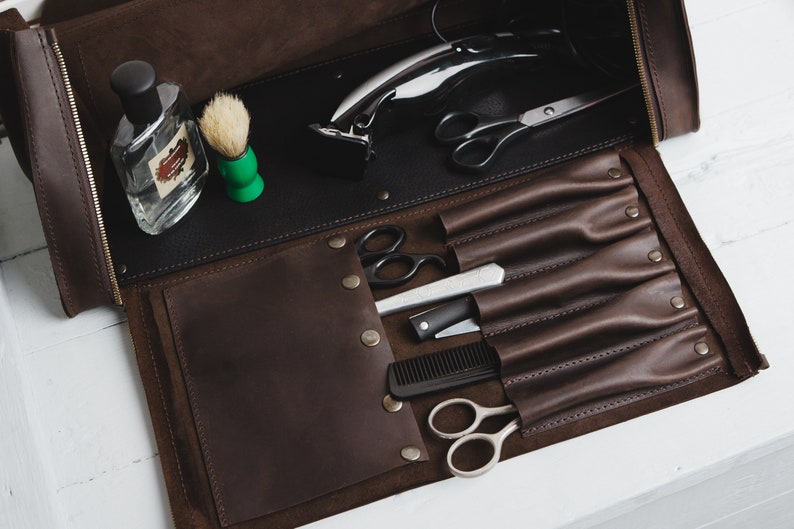 Op de afbeelding: Open bruin leren barbierkit met een zwart interieur. De kit bevat een flesje parfum, een scheerkwast, een elektrische tondeuse, een schaar, een recht scheermes, een kam en andere verzorgingsgereedschappen. Op een wit oppervlak.
