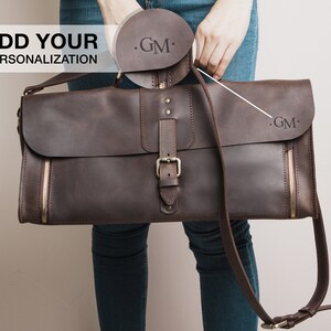 Op de afbeelding: Een bruine leren messenger bag met een klepsluiting, gesp en ritssluiting details. De tas heeft een schouderriem en heeft de initialen "GM" op de flap. De tekst "ADD YOUR PERSONALIZATION" is ook zichtbaar.
