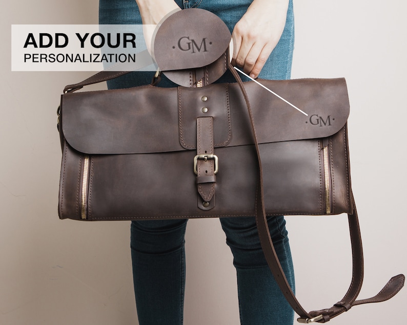 Op de afbeelding: Een bruine leren messenger bag met een klepsluiting, gesp en ritssluiting details. De tas heeft een schouderriem en heeft de initialen "GM" op de flap. De tekst "ADD YOUR PERSONALIZATION" is ook zichtbaar.
