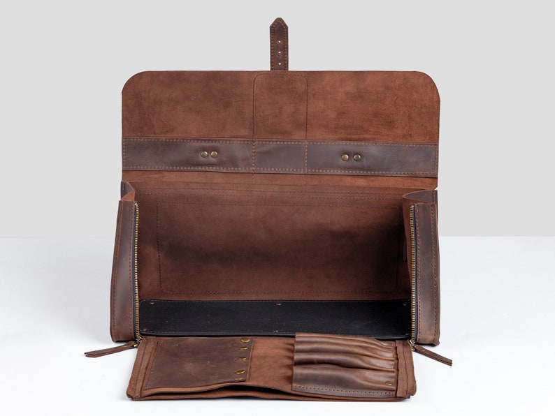 Op de afbeelding: Open bruinleren etui met ritssluiting en meerdere compartimenten. De etui heeft een flap met een gespsluiting en een gedeelte voor pennen of potloden. De binnenkant is gevoerd met zwarte stof.
