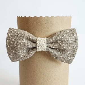May include: A brown bow tie with white polka dots and a cream-colored center knot is displayed on a brown paper tube. The bow tie is made of a textured fabric.