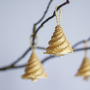 May include: Three straw Christmas tree decorations hanging from bare branches. The decorations are made of woven straw and are a light brown colour.