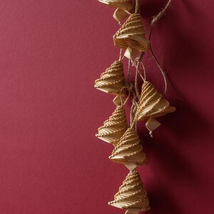 May include: A string of handmade Christmas tree ornaments. Each tree is constructed from thin, light-colored wooden sticks, arranged in a spiral pattern. The ornaments hang from a natural twine against a deep red background.