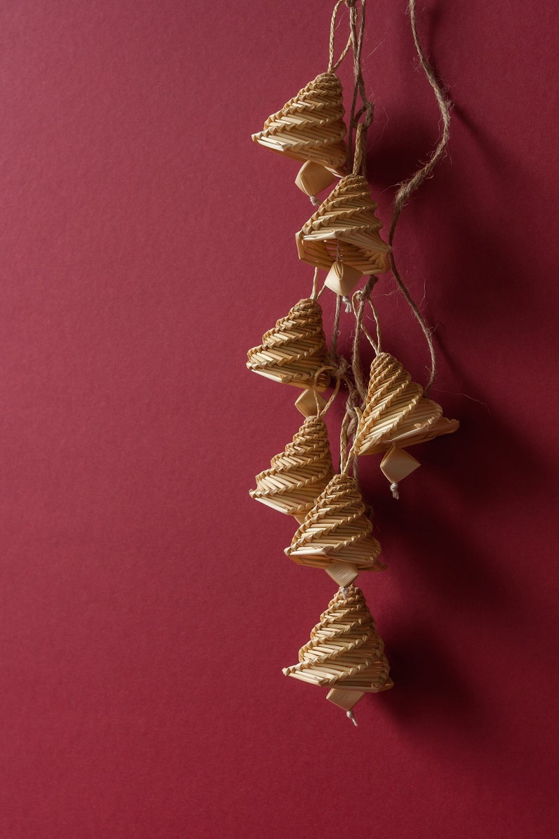 May include: A string of six woven straw bells hanging against a red background. The bells are all the same shape and size, and they are made of a light brown straw.