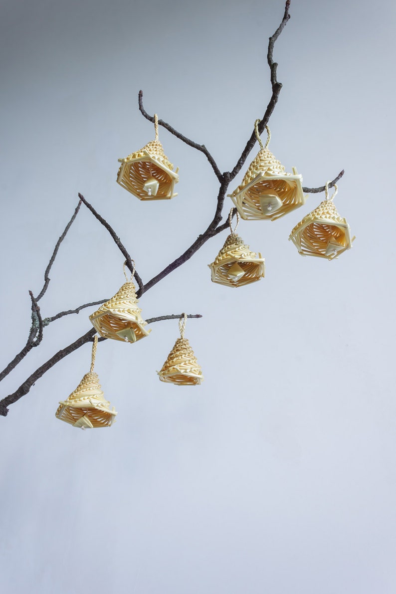 May include: A collection of eight woven straw ornaments hanging from a bare branch. The ornaments are shaped like bells and are a light beige colour.