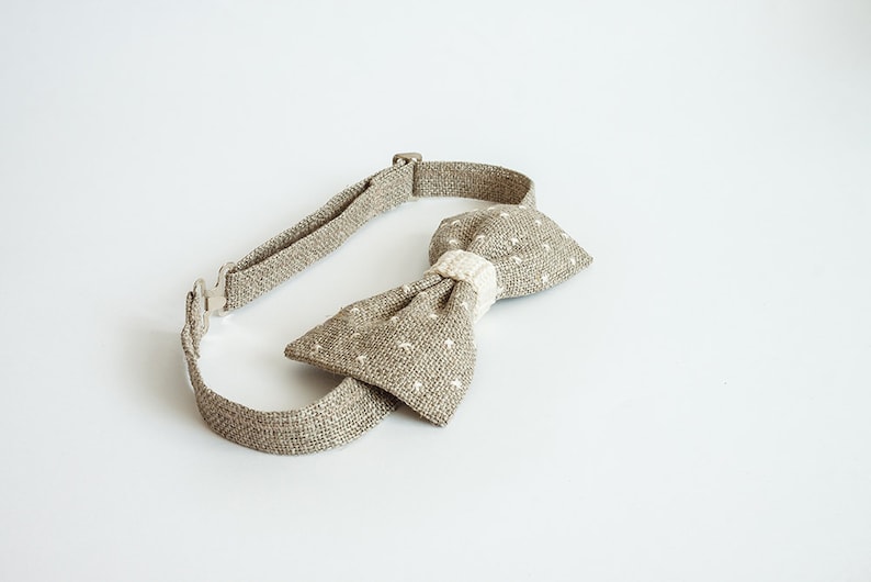May include: A tan bow tie with a white dotted pattern and a cream-colored center. The adjustable neckband is made of the same tan fabric. The bow tie is a classic accessory for formal or semi-formal attire.