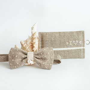 May include: A tan bow tie with white polka dots and a matching wallet. The bow tie has a cream-colored center and is next to a seashell. The wallet has the initials "IV" embroidered on it.