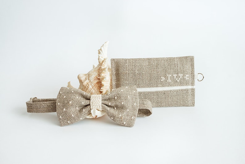 May include: A tan bow tie with white polka dots and a matching wallet. The bow tie has a cream-colored center and is next to a seashell. The wallet has the initials "IV" embroidered on it.