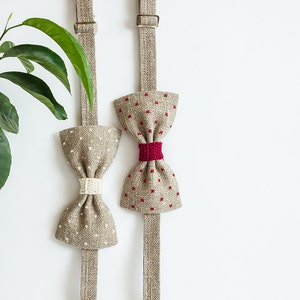 May include: Two bow ties with adjustable straps hang against a white background. One is beige with white dots and a cream-colored center. The other is beige with red squares and a red center. A green plant is in the upper left corner.