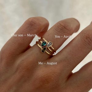 Puede incluir: Tres anillos de tono dorado, cada uno con una piedra preciosa en forma de pera. Las piedras son azul oscuro, transparente y verde oliva. El texto en la imagen dice: Craft your story with our iconic birthstone rings, Our son – March, You – April, Me – August.