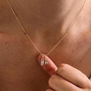 Op de afbeelding: Een gouden ketting met twee kleine hangers: een rechthoekige, heldere steen in baguette-slijpvorm en een peervormige, lichtgroene steen. Beide bedels hangen aan de fijne gouden ketting met behulp van kleine ronde ringetjes.