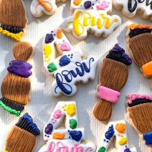 May include: A set of nine decorated sugar cookies featuring the number four and the word "four" in various colours and styles. The cookies are decorated with paint splatters and paintbrushes.