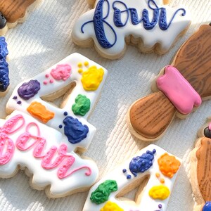 May include: Four decorated sugar cookies with colourful paint splatter designs. The cookies are shaped like the number four and the word "four".