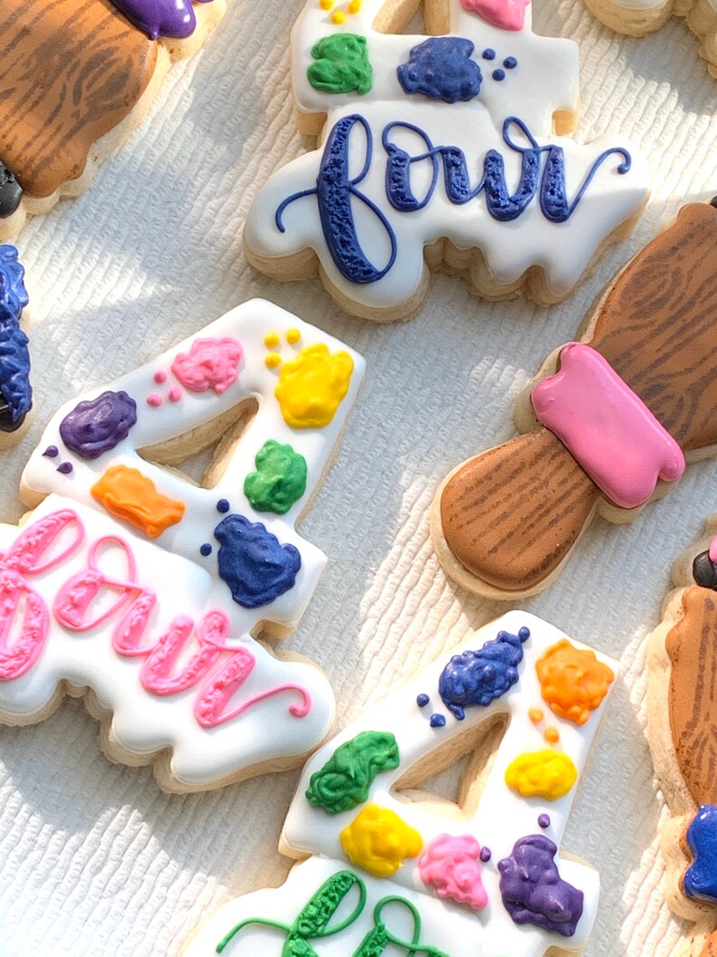 May include: Four decorated sugar cookies with colourful paint splatter designs. The cookies are shaped like the number four and the word "four".