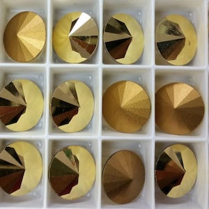 May include: A close-up of a container of 18 gold-colored round rhinestones. The rhinestones are arranged in three rows of six. Each rhinestone has a pointed top.