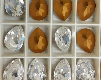 May include: A collection of 10 teardrop-shaped glass rhinestones in a clear and gold colour. The rhinestones are arranged in a grid pattern in a white tray.