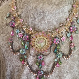 May include: A multi-layered necklace with a large, ornate, gold-toned pendant featuring a floral design. The necklace is made of various beads, crystals, and flowers in shades of pink, green, and blue.