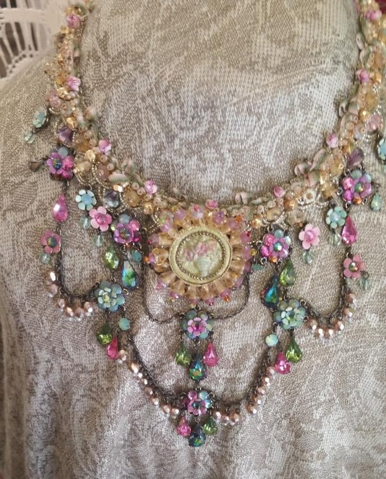 May include: A multi-layered necklace with a large, ornate, gold-toned pendant featuring a floral design. The necklace is made of various beads, crystals, and flowers in shades of pink, green, and blue.