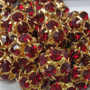 May include: A close-up of a pile of red rhinestone beads with gold settings. The beads are round and have a faceted design.
