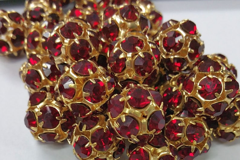 May include: A close-up of a pile of red rhinestone beads with gold settings. The beads are round and have a faceted design.
