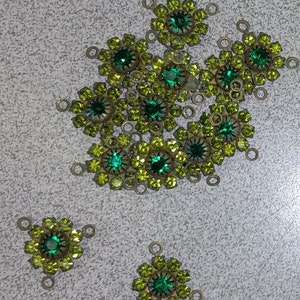 May include: A group of 14 green and gold metal embellishments with a flower design and a green gem in the center. Each embellishment has three loops on the back for attaching to fabric.