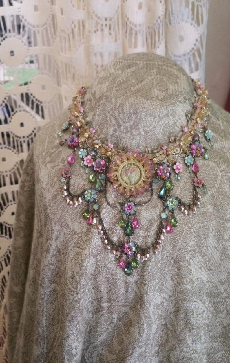 May include: A delicate, multi-layered necklace with a floral design. The necklace features pink, green, and gold beads, as well as small flower accents. The centre of the necklace features a large, round, gold-toned pendant with a floral design.