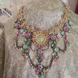 May include: A multi-layered necklace with a large, ornate centrepiece. The necklace is made of gold-toned metal and features pink, green, and blue crystals, as well as small flowers.